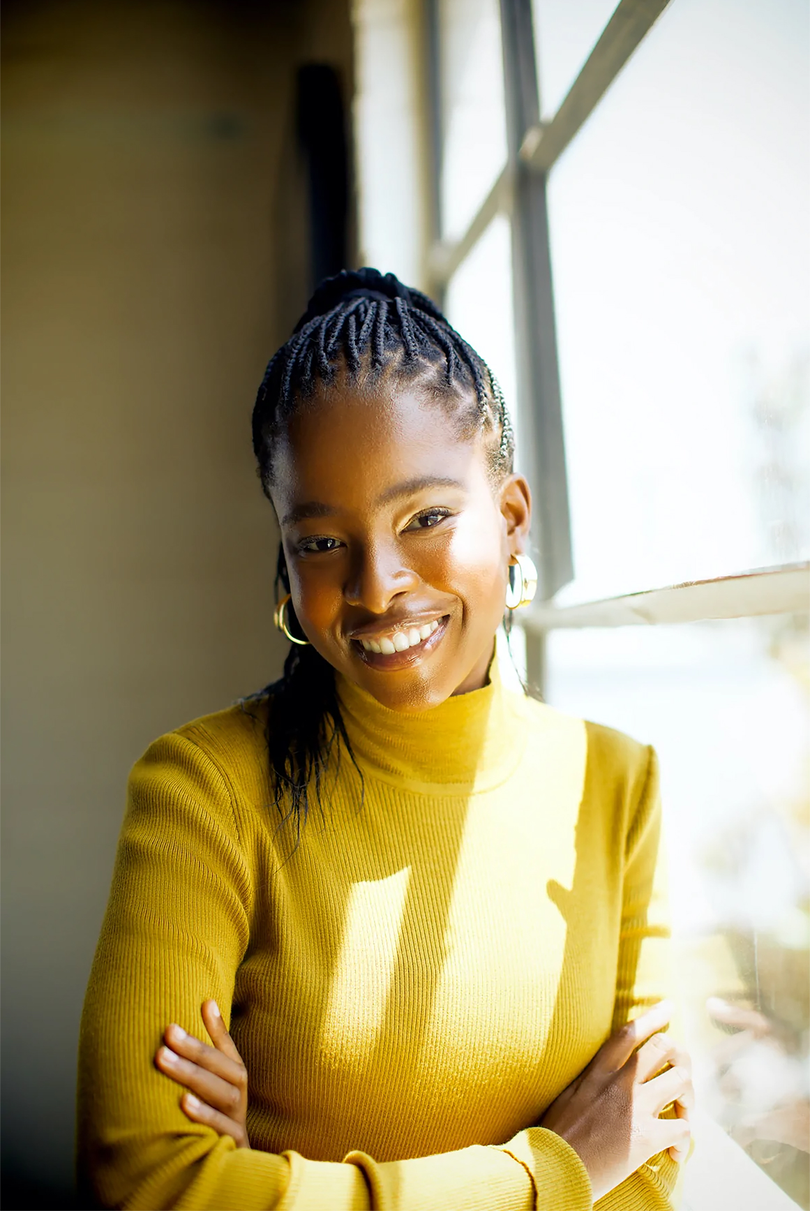 A smiling young woman sitting next to a window; she is bathed in sunlight